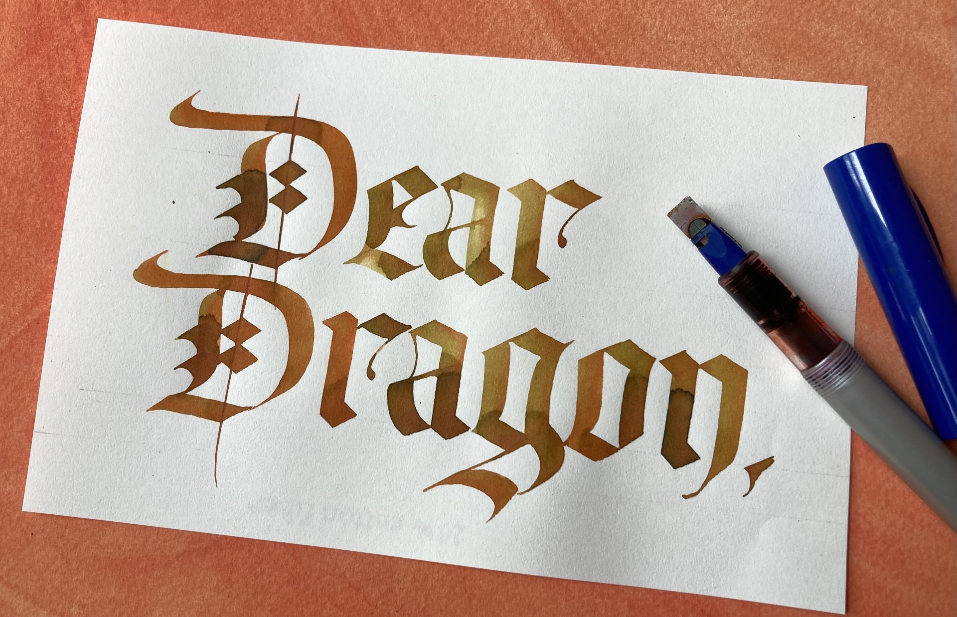 A calligraphy piece with the words Dear Dragon, written on a piece of paper with a broad edge nib pen next to it.