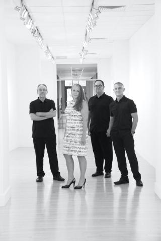 a black and white image of a group of four people, three male and one female. They are in a long hallway that is completely white. The female is in front and wearing a dress of musical symbols and lines. the men are dressed in black and are in relaxed positions.