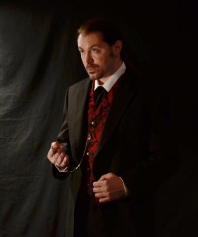 A man dressed in a suit with a red and black undershirt. The man is looking to the left and standing. 